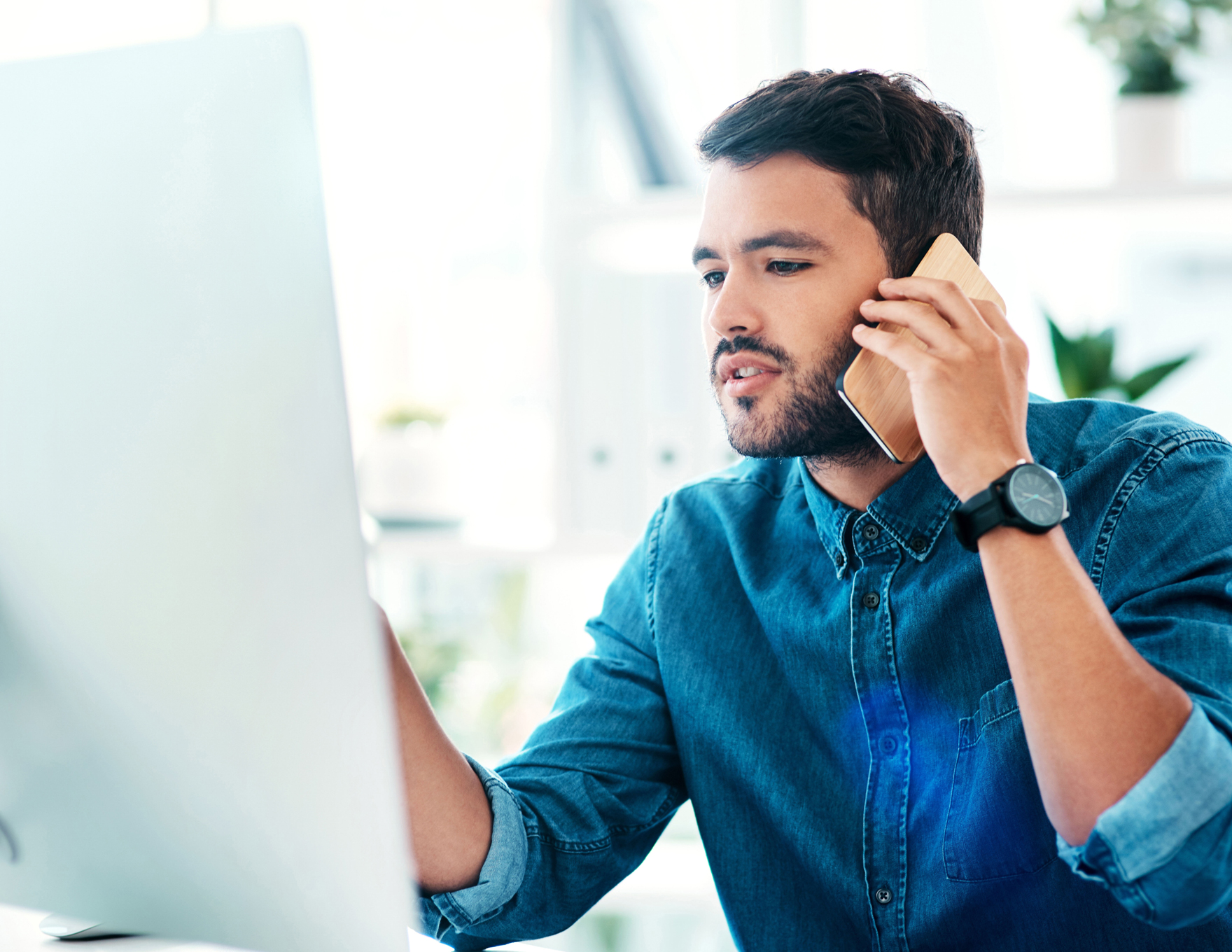 Man in a denim shirt speaks on a smartphone at a bright desk setup with a large monitor nearby.