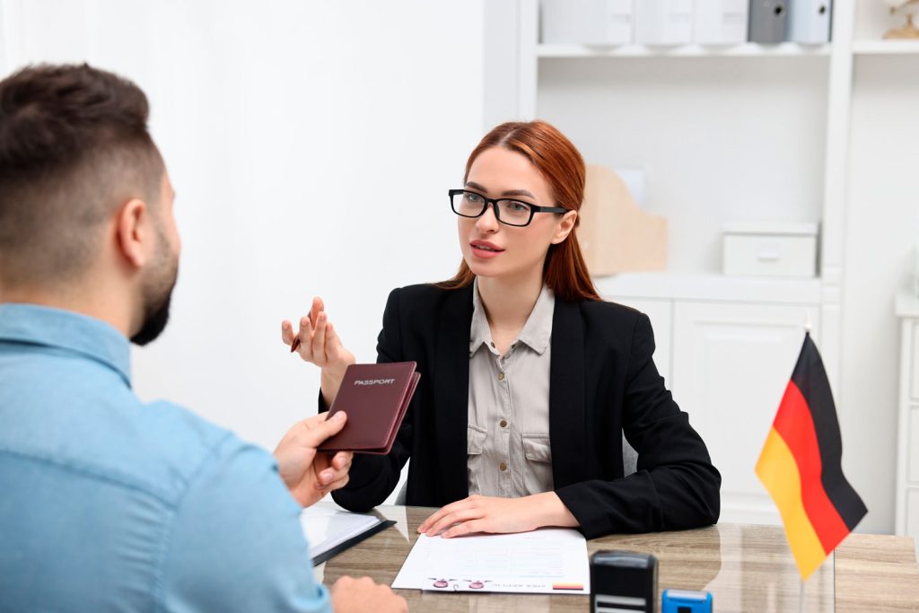 Woman in blazer speaks with a man across a desk; she holds a passport and they discuss documents in an office setting with a German flag on the desk edge.